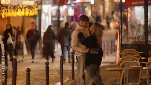 Canvas Print A romantic couple embraces on a Parisian street at night surrounded by tables and festive Christmas lights