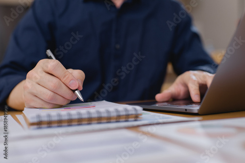 businessman writing in notebook while using laptop, taking notes from financial data and business reports in office environment. Productivity and planning concept.