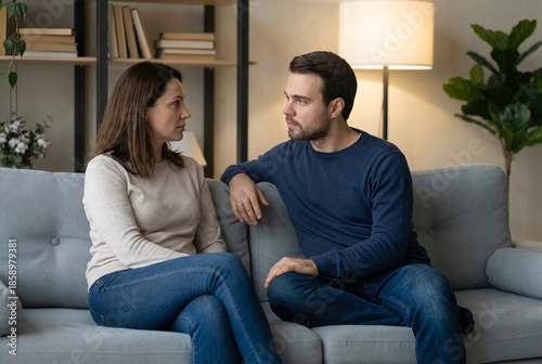 Serious couple sitting on sofa at home having difficult conversation, man and woman discussing relationship problems, conflict resolution or financial planning in modern living room.