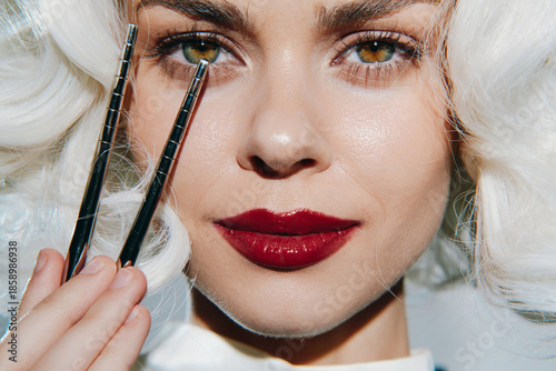 Tableau sur toile Elegant woman with striking makeup holding tweezers, showcasing beauty artistry and vibrant emotions against a minimalist background