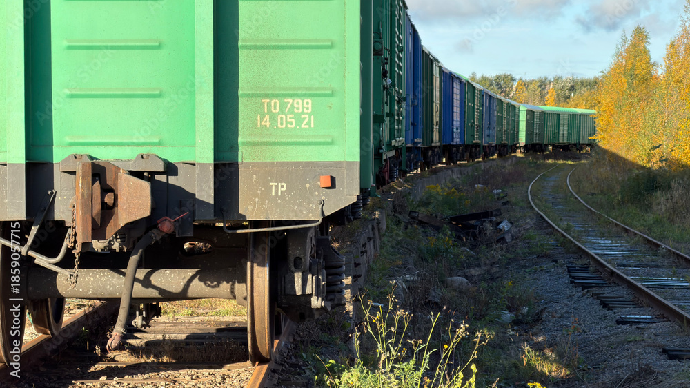 Obraz premium A close-up view of a green freight train car on tracks, with a long line of train cars curving in the background, surrounded by autumn foliage