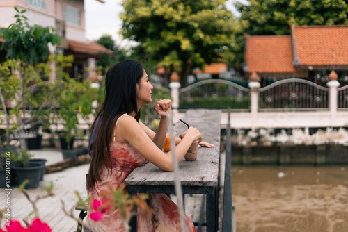 Schilderij op canvas Young asian woman enjoying a drink at a canalside cafe, a moment of relaxation