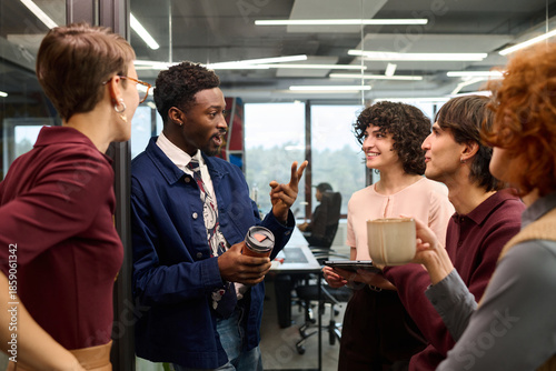 Group of young adult multiethnic colleagues standing in modern office, engaging in lively conversation, Black man gesturing while holding reusable coffee cup, others listening and smiling