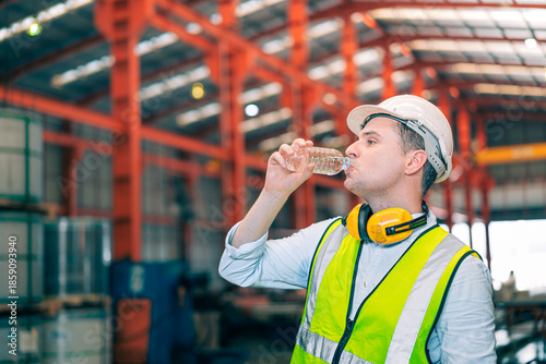 Thirsty Caucasian industrial engineer man wear safety helmet vest drinking water from plastic bottle in factory. Tired worker taking break refreshment. Manufacturing plant hydration health care