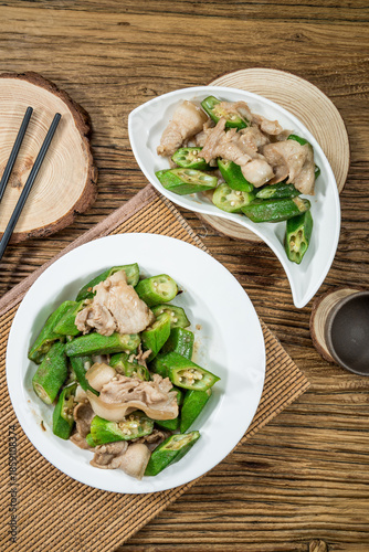 Two plates of stir-fried pork slices with fresh green okra and garlic, served on a wooden table with chopsticks for a rustic presentation.