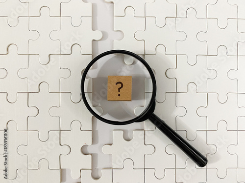 Foto Magnifying glass focuses on a cardboard question mark block atop white jigsaw pi