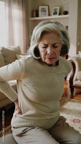 Elderly woman experiencing back pain while sitting on a sofa at home, vertical shot