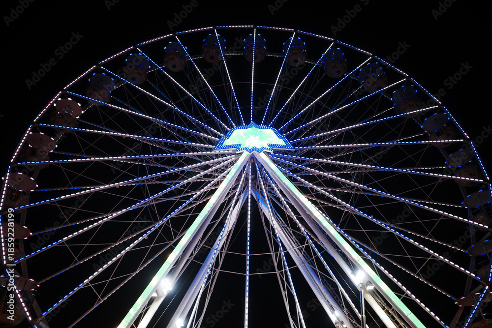 Fototapeta premium Low-angle night photograph of a large illuminated Ferris wheel against a deep black sky.