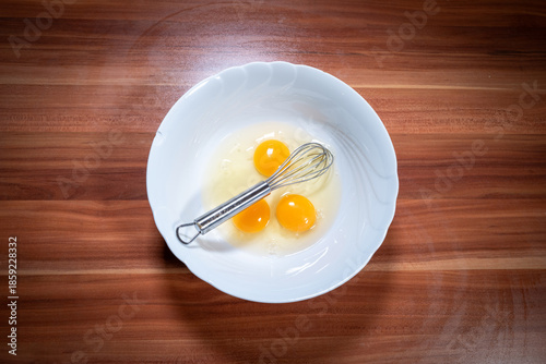 Top-down food photography showing three raw eggs cracked into a white ceramic bowl with a metal whisk, placed on a wooden kitchen countertop