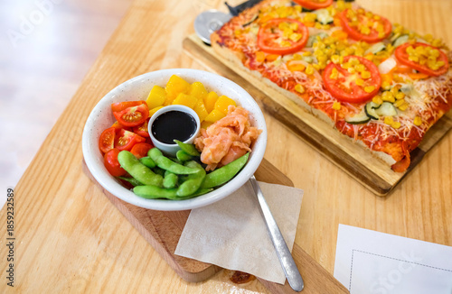 Top view of poke bowl with fresh salmon, mango, edamame, cherry tomatoes and soy sauce, serving on restaurant table next to vegetarian focaccia, in casual lunch. Organic and natural fast food concept