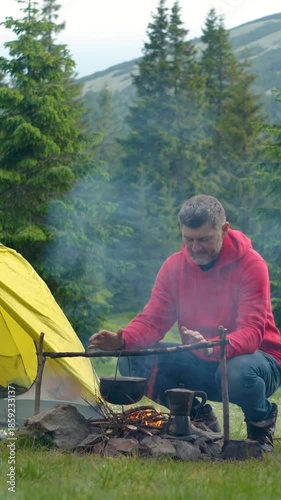 Man Cooking Food On Campfire Near Tent In Mountain Forest, Camper preparing meal on open fire beside tent in green mountain landscape