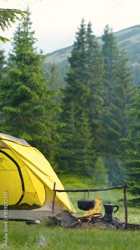 Yellow Camping Tent Beside Fire With Cooking Pot In Mountain Forest, Peaceful outdoor campsite scene showing wilderness breakfast preparation at dawn