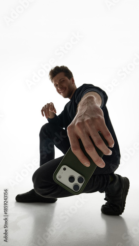Man crouching on floor while holding smartphone in dynamic pose on white. Concept of youth focused smartphone advertising, lifestyle brand visuals, social media marketing, and mobile product promotion