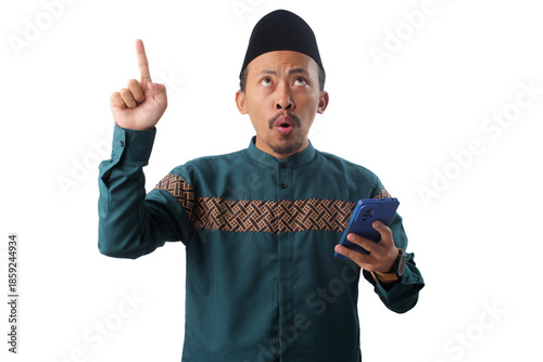 Asian Muslim man in a green traditional outfit holding a smartphone and pointing at empty copy space, isolated on white background. Perfect for promotional or communication themes
