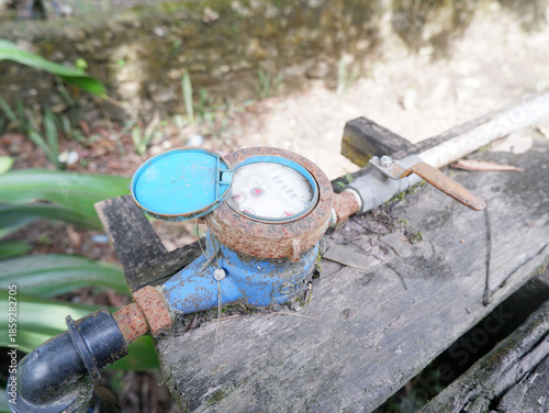 ​Close-up of a residential water meter with a blue lid and valve on wooden background. Concept of water consumption, utility management, and plumbing system in a home garden