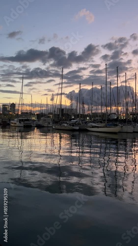 Sunset marina with sailboats and yachts reflected on calm water under dramatic cloudy sky, peaceful harbor landscape at dusk with moored boats, nautical atmosphere and cinematic travel background