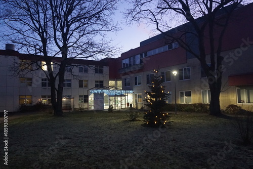 Beleuchtetes Krankenhausgeländer mit Christbaum auf Wiese bei Frost und Kälte in der Nacht im Winter