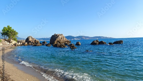 Mountains and a port are visible in the hot haze on the seashore. Near the pebble shore with pine trees, large stones lie in the clear water. The waves are foaming. Sunny autumn weather and blue sky