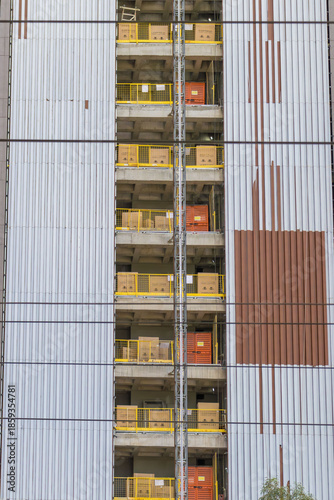 High-rise building under construction with exposed concrete floors, yellow safety railings, and metal facade panels.