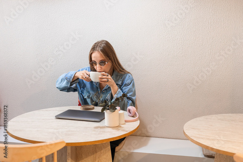 A young Caucasian woman with long brown hair sits at a wooden table, sipping coffee from a white cup. A laptop and a small plant are on the table.