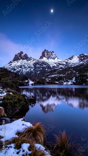 A serene winter night landscape featuring snowy mountains reflecting in a calm lake under a star-filled blue sky