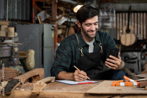 Carpenter using smartphone and taking notes in woodworking workshop, small business craftsman
