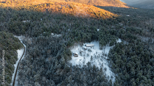 An aerial winter landscape of the Adirondack Mountains at sunrise. Golden light hits the snowy peaks above a frost-covered evergreen forest.