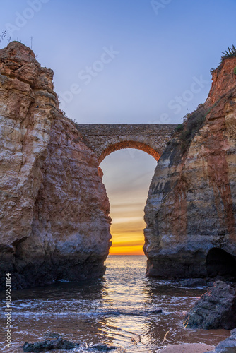 A dramatic vertical shot of the famous stone arch bridge connecting two sandstone cliffs at Students Beach (Praia dos Estudantes) during a vibrant sunrise.