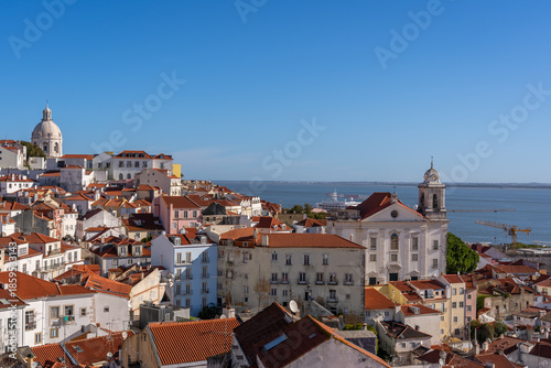 High-angle view from Miradouro do Recolhimento showing the red-tiled rooftops of Lisbon, the Church of Santo Estevao, the National Pantheon dome, and the Tagus River under a clear blue sky.