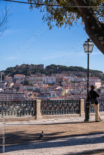 A male traveler standing at a viewpoint (Miradouro) looking over the historic city of Lisbon towards the castle. Lifestyle travel concept with a scenic urban background.