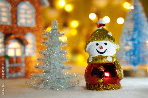 A snowman between a white and blue Christmas tree with an orange house on a blurred background with yellow lights