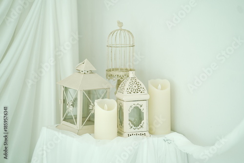 Elegant Home Decor with Lanterns, Candles, and Birdcage Arranged on White Fabric Background for Cozy and Warm Interior Atmosphere