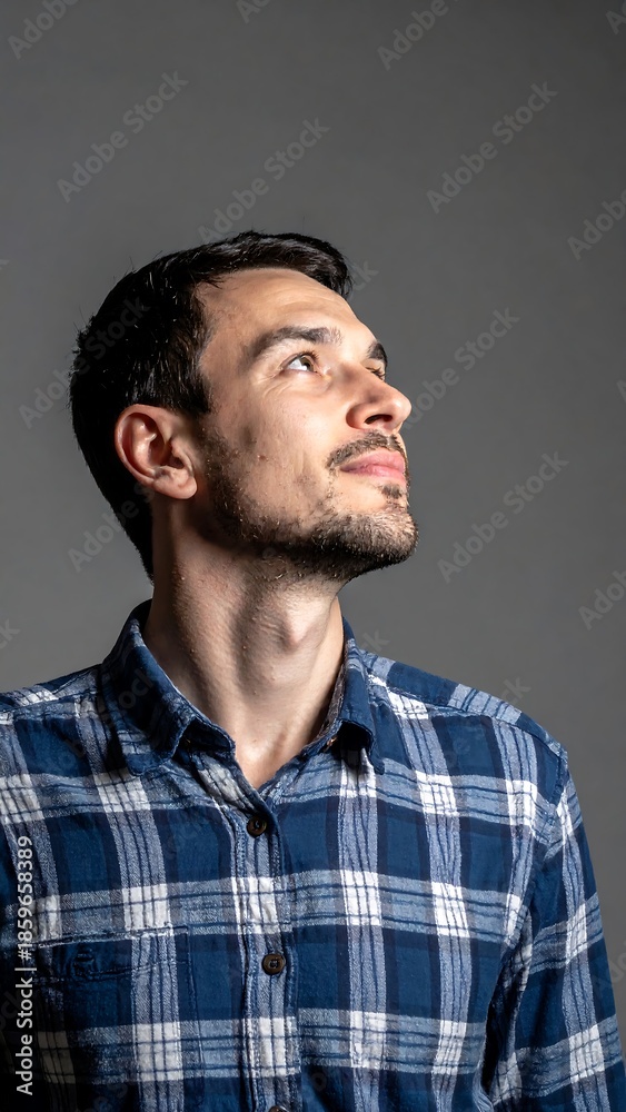 Fototapeta premium Man gazes upward in a blue plaid shirt against a plain grey backdrop, studio lighting illuminates his face
