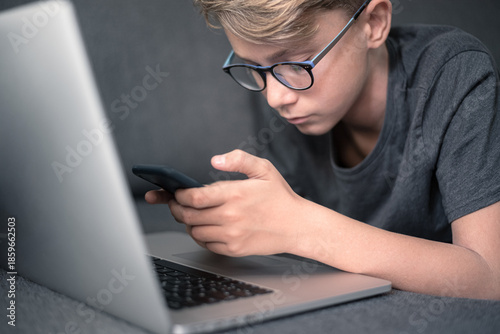 A young Caucasian boy wearing glasses lies on a grey sofa using a smartphone while an open laptop sits nearby illustrating the concept of modern digital multitasking and student distraction