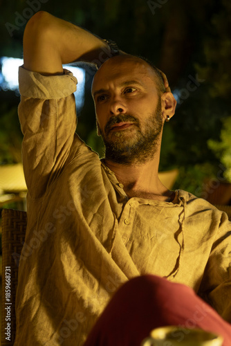 A portrait of a mature man sitting at a cozy outdoor cafe table during a warm summer evening, enjoying a cup of coffee. He looks relaxed and thoughtful in a modern urban lifestyle setting.