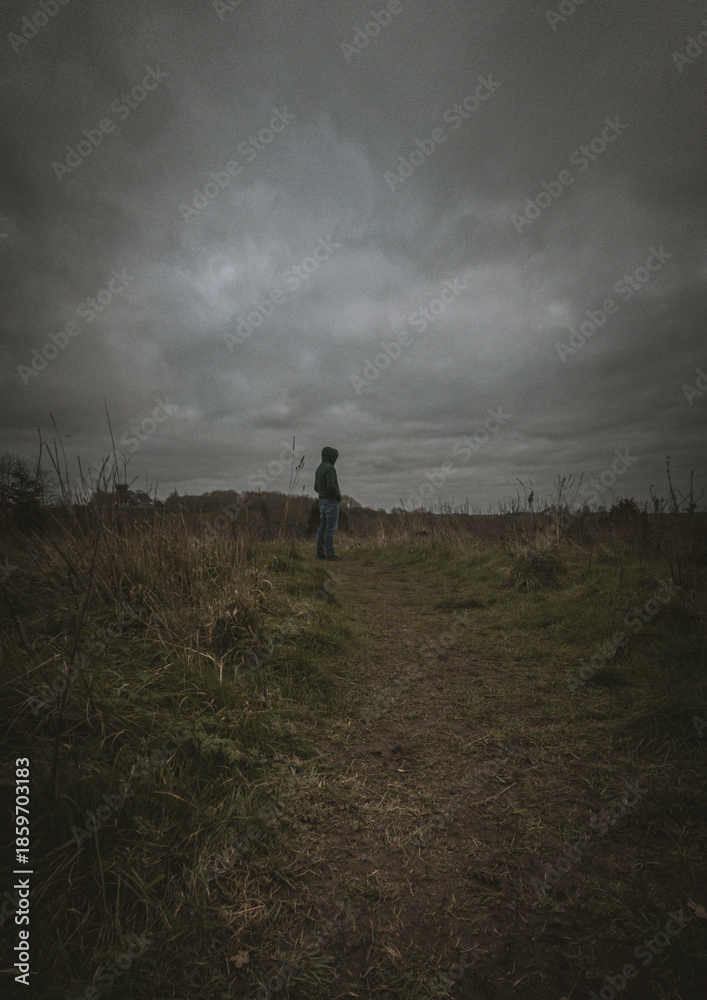 Fototapeta premium Lonely man in a hoodie standing on a meadow under a dramatic cloudy sky, moody atmosphere, solitude concept.