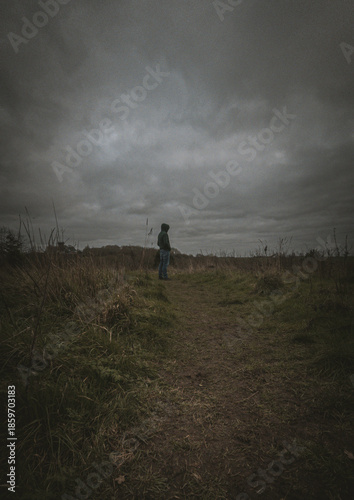 Lonely man in a hoodie standing on a meadow under a dramatic cloudy sky, moody atmosphere, solitude concept.