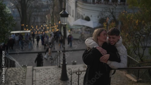 A romantic couple embraces in Montmartre Paris France The woman kisses the man on the cheek The background is blurred showing the Sacre-Coeur basilica and a bustling street scene.