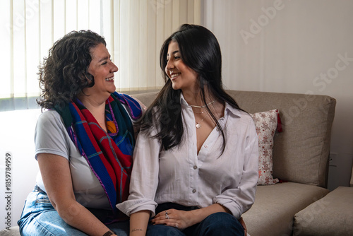 Happy Hispanic mom and adolescent girl sitting on a sofa facing each other, smiling and talking. Concept of family communication, trust, and mother-daughter bonding.