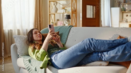 Relaxed girl watching smartphone screen lying comfortable couch. Smiling young