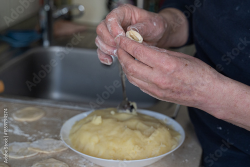 Elderly hands spoon mashed potato filling onto dough circle beside white bowl in soft focus, authentic cooking at home scene capturing traditional dumpling preparation process.