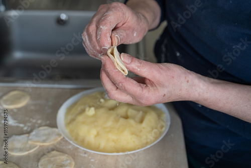 Elderly hands spoon mashed potato filling onto dough circle beside white bowl in soft focus, authentic cooking at home scene capturing traditional dumpling preparation process.