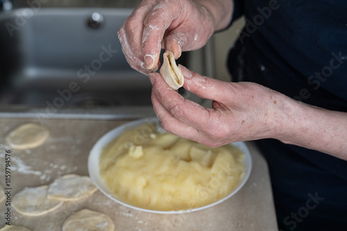 Elderly hands spoon mashed potato filling onto dough circle beside white bowl in soft focus, authentic cooking at home scene capturing traditional dumpling preparation process.