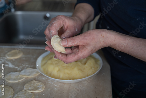 Elderly hands spoon mashed potato filling onto dough circle beside white bowl in soft focus, authentic cooking at home scene capturing traditional dumpling preparation process.