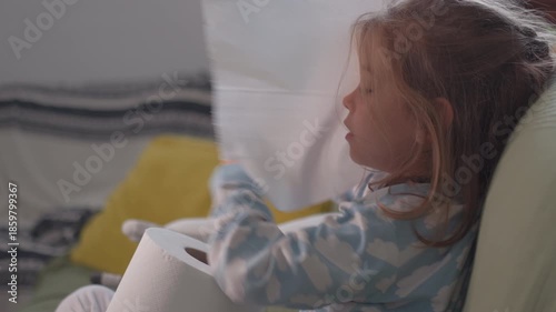 Side view of young sick girl blowing nose into tissue sitting on sofa in living room. Female child suffering from cold and flu at home at night