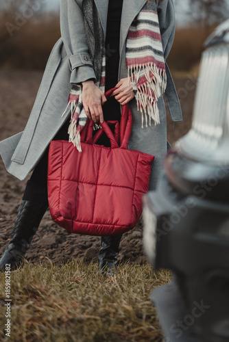 Stylish woman in a gray coat and striped scarf holding a red quilted puffer bag against a dark field, vertical shot in natural light