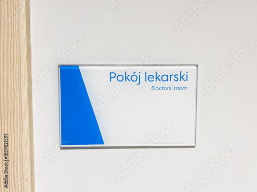 A horizontal photo showing a modern information plate with the words Doctor's Room and Doctors' room, mounted on a white wall next to a wooden frame in a bright, sterile interior of a medical clinic w