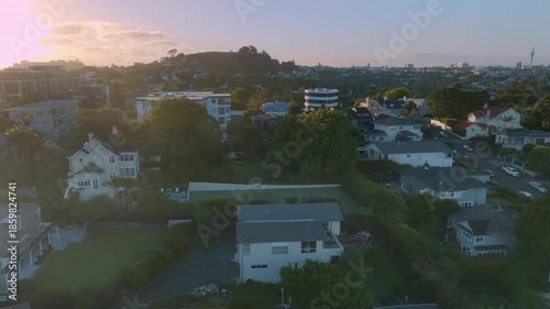 Aerial view of Remuera, Auckland, New Zealand, showcasing a residential neighborhood with houses, trees, and a distant city skyline at sunset. The scene captures the urban landscape.