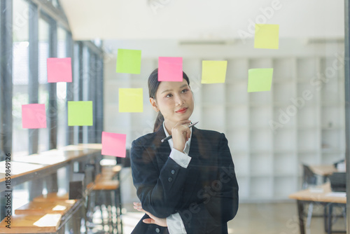 Bild auf Leinwand Thoughtful asian woman with hand on chin looking at sticky notes on glass wall,