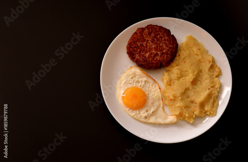 Beefsteak with fried egg and mashed potatoes in a round ceramic plate on a black background.